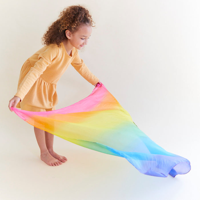 A young girl with curly hair, barefoot and in a yellow dress, stands against a light background, captivated by an Enchanted Playsilk from Sarahs Silks. She spreads it out before her, exploring its creative potential for endless open-ended play possibilities.