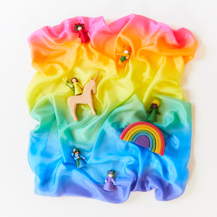 Sarahs Silks Enchanted Playsilk creates a colorful backdrop for small toys. A wooden unicorn and vibrant figures, placed on the mulberry silk fabric, inspire open-ended, imaginative play.