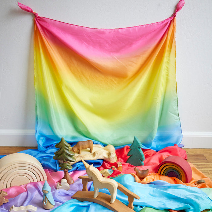 A vibrant Enchanted Playsilk by Sarahs Silks, featuring a mulberry silk gradient of pink, yellow, and blue, adorns the wall. Wooden toys such as trees, animals, and a rainbow encourage open-ended play among the crumpled pink, red, and blue fabric.