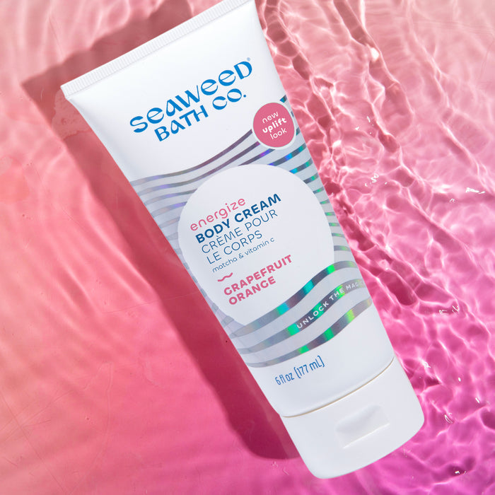 A tube of The Seaweed Bath Co. Energizing Body Cream, enriched with seaweed and labeled with grapefruit and orange, floats on pink water, featuring a new uplift note and the phrase unlock the magic.