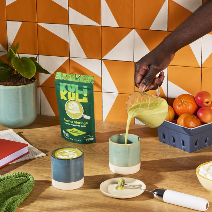 A hand pours Kuli Kuli Foods Energizing Organic Superfood Latte, a green moringa drink, into a mug on a wooden table with another mug, its pouch, citrus fruits, books, and a matcha whisk. Orange geometric tiles add vibrancy. Rich in B vitamins.