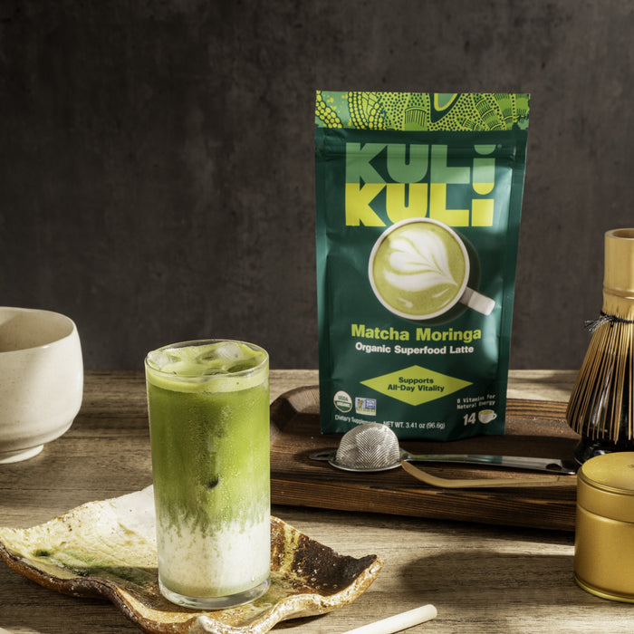 A glass of iced Energizing Organic Superfood Latte by Kuli Kuli Foods sits on a ceramic plate. Behind it stands the product’s green package, highlighting natural B vitamins. Tea-making utensils are arranged on a wooden tray nearby.