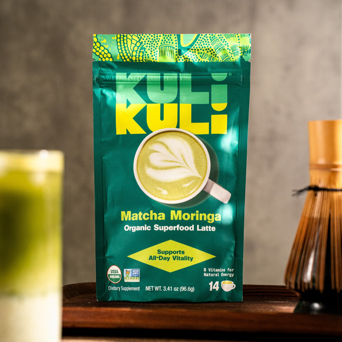 A green pouch labeled Kuli Kuli Foods Energizing Organic Superfood Latte stands upright on a table, featuring B vitamin-rich moringa and matcha, with a green latte, bamboo whisk, and softly blurred background beside it.