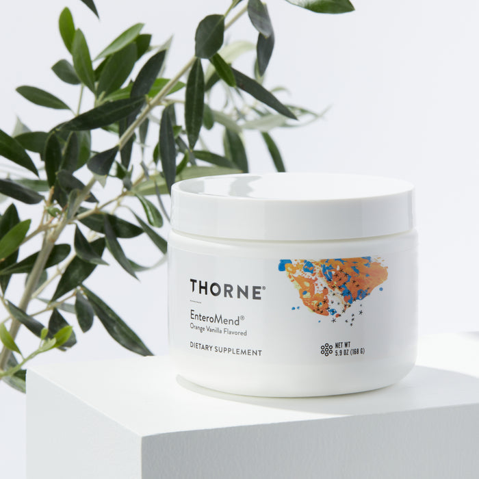 A Thorne EnteroMend white container, supporting intestinal health, is on a white surface. The orange vanilla flavor enhances the green leaves and branches against a light backdrop, adding a natural touch.