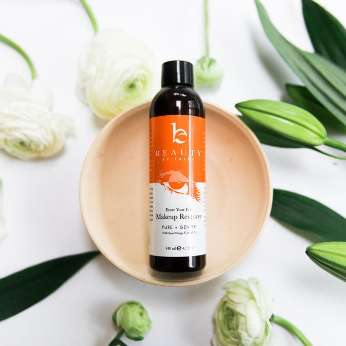 A dark bottle of Beauty by Earths Erase Your Face Makeup Remover, featuring an orange label with white text and a crab illustration, is surrounded by white flowers and green leaves on a light wooden tray. It is made from organic ingredients for naturally healthy skin.