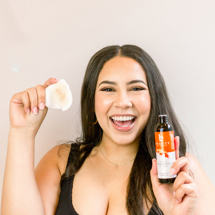 A person with long dark hair beams holding a used cotton pad and Beauty by Earths Erase Your Face Makeup Remover. Their naturally radiant skin shines, highlighted by a black top against a plain background.