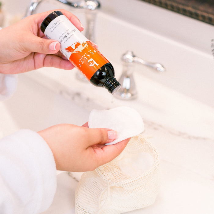 Someone in a white robe pours Beauty by Earths Erase Your Face Makeup Remover onto a white cotton pad. The cotton pad is held over a mesh bag by a white sink with a silver faucet, and the brown bottle features an orange label, suggesting its likely filled with organic ingredients.