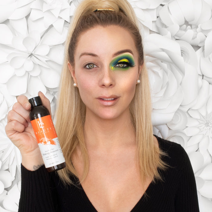 A woman with a blonde ponytail holds a dark bottle of Beauty by Earths Erase Your Face Makeup Remover. One side of her face is boldly made up, and the other glows naturally. She wears a black top against a textured floral white background.