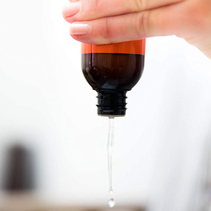 A hand holds an upside-down amber glass bottle of Beauty by Earths Erase Your Face Makeup Remover, featuring a red label. A few drops are dispensed, made with organic ingredients to naturally promote healthy skin against a blurred background.