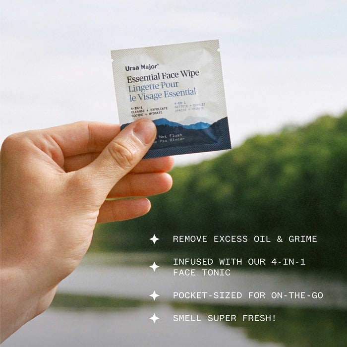 A hand holds a packet of Ursa Major Essential Face Wipes, Individually Wrapped, outdoors with blurred trees and water in the background. Text highlights benefits: removes excess oil, 4-in-1 tonic, pocket-sized, and fresh scent.