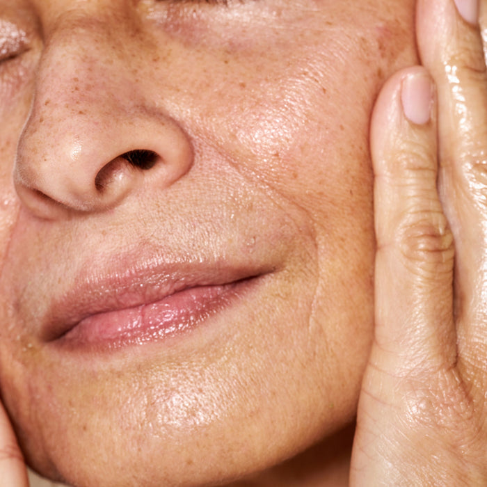A close-up of a persons freckled, smooth face with eyes closed reveals their glowing complexion. Hands gently caress the cheeks, indicating a dewy look from OSEAs nourishing Essential Hydrating Oil application.
