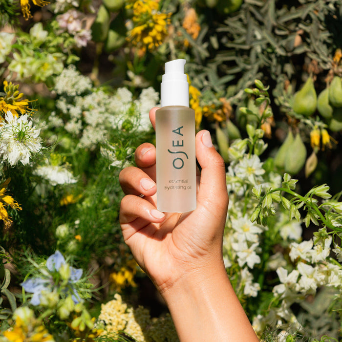 A hand holds a translucent OSEA Essential Hydrating Oil bottle with a white pump amidst greenery and flowers, accentuating its antioxidant-rich oils for luminous skin.