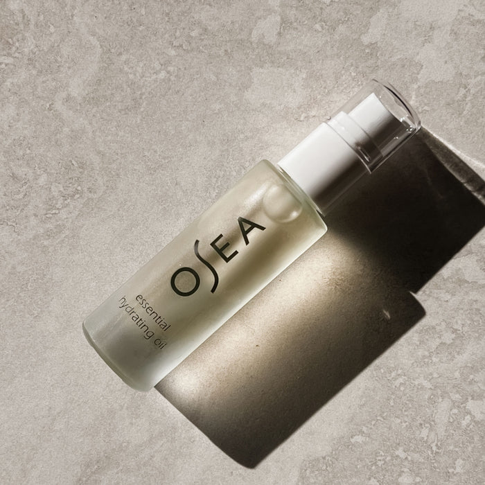 A frosted bottle of OSEAs Essential Hydrating Oil rests on a textured surface, casting a shadow. The label shows the brand and product details in black text. This anti-aging face oil, with a white pump dispenser, is designed to reduce fine lines and enhance skin glow.