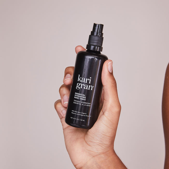 A hand holds a black spray bottle of Kari Gran Essential Hydrating Tonic against a neutral background. The bottle features white text with brand and product info, perfect for rehydrating skin and supporting the moisture barrier.