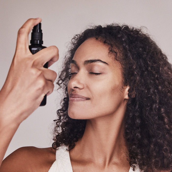 A woman with curly hair, wearing a white top, smiles with her eyes closed while spraying Kari Grans Essential Hydrating Tonic from a black bottle near her face against a neutral background. This tonic rehydrates skin and supports the moisture barrier.