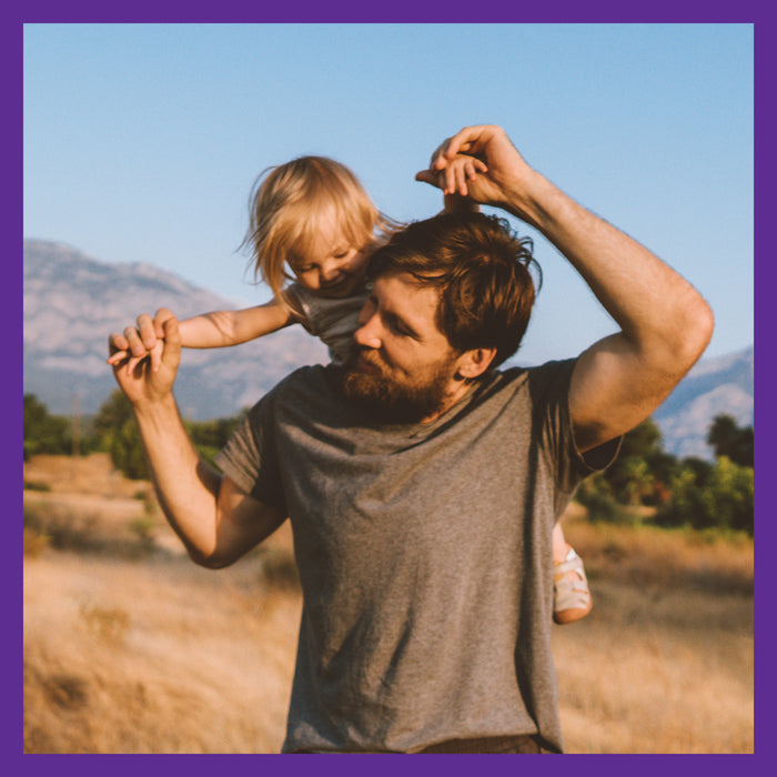 A bearded man in a gray t-shirt carries a child on his shoulders, playfully leaning forward amidst green grass and mountains bordered in purple. He radiates vitality—maybe thanks to New Chapters Every Man™s One Daily 40+, a whole-food fermented multivitamin for men over 40.