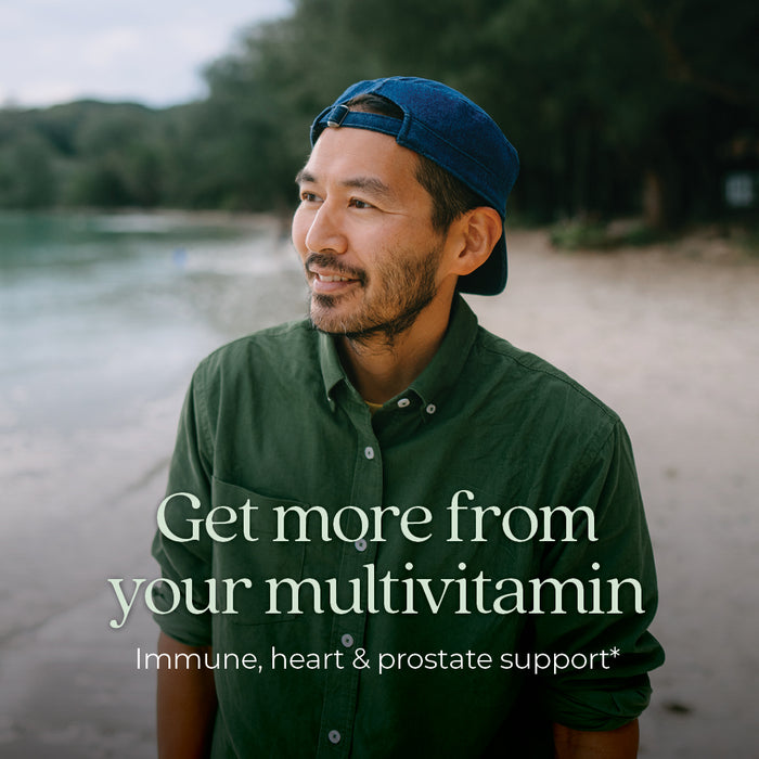Man in a green shirt and blue cap smiles while standing on a beach. Text on the image reads, "Get more from your multivitamin: Immune, heart &amp; prostate support." Lush trees and calm water are in the background.