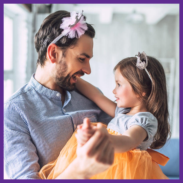 A man and a young girl smile at each other as he playfully wears her pink hair accessory. She twirls in an orange tutu around him in their loving home, a scene filled with joy and affection, no New Chapters Every Man™s One Daily Multivitamin for energy or stress support needed.
