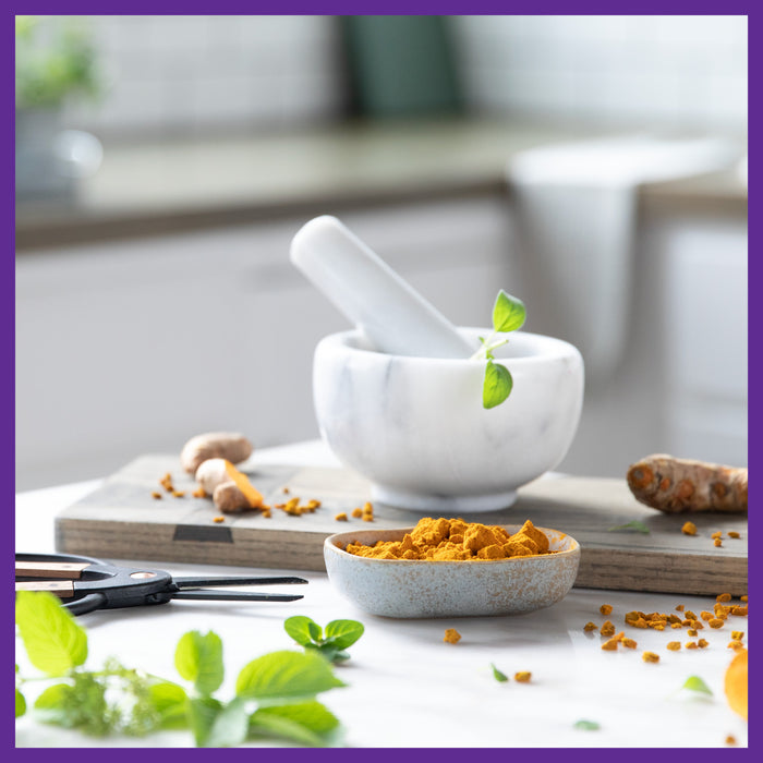 A marble mortar and pestle grace the kitchen counter with turmeric, mint leaves, and scissors, forming a vibrant setup with organic ingredients that complements New Chapters Every Man™s One Daily Multivitamin for a refreshing workspace supporting immune health.