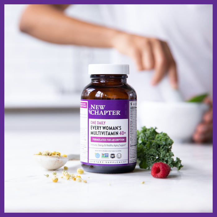 A bottle of New Chapters Every Woman™s One Daily 40+ Multivitamin sits amid fresh kale and a raspberry, offering essential vitamins and minerals for energy support, as a person prepares food in the bright white kitchen.