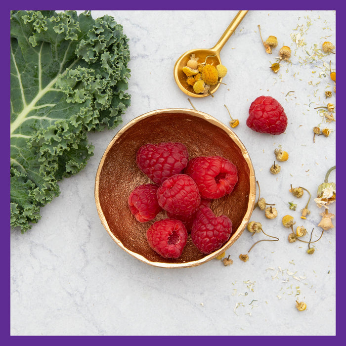 A small golden bowl with raspberries rests on a marble surface, offering a touch of energy support from New Chapter Every Womans One Daily 40+ Multivitamin. Loose chamomile flowers and a gold spoon with dried chamomile surround it, while a large kale leaf lies to the left, framed by a purple border.