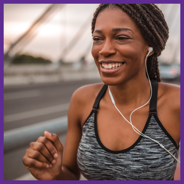 A woman radiates vibrant energy while jogging on a bridge at sunset, wearing a gray tank top and earphones. The blurred background mimics her lively stride, as if fueled by New Chapters Every Woman™s One Daily 40+ multivitamin.