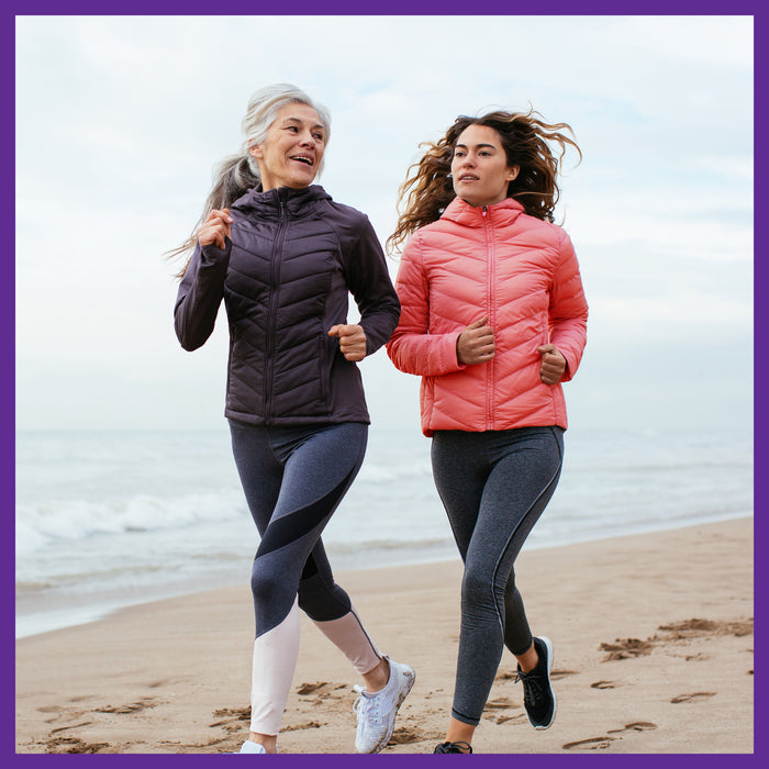 Two women jog on a sandy beach, embracing healthy aging with the refreshing ocean waves. They wear dark purple and pink leggings and insulated jackets under an overcast sky, supported by New Chapters Every Woman™s One Daily 55+ Multivitamin for cellular energy, heart & immune support.