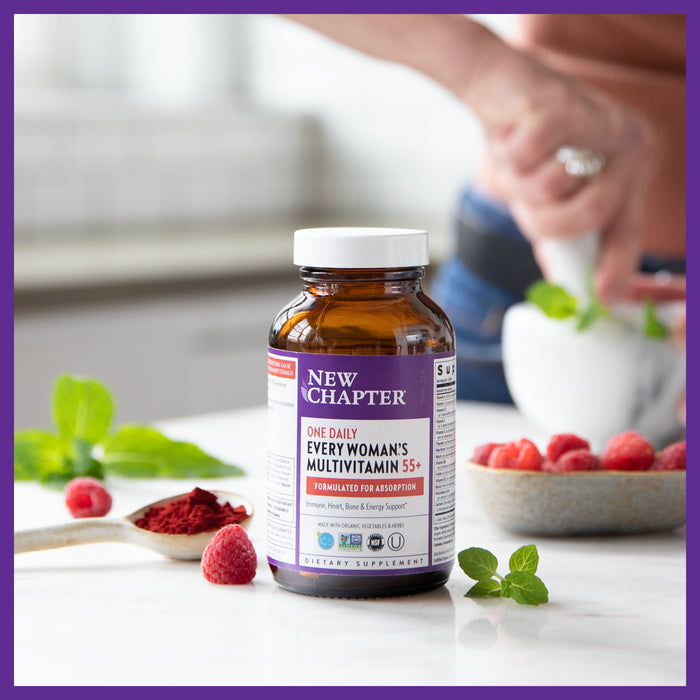 A New Chapter Every Woman’s One Daily 55+ Multivitamin bottle rests on a kitchen counter with raspberries, a small bowl of red powder, and mint leaves held by someone in the background. The label promotes cellular energy and heart and immune support.
