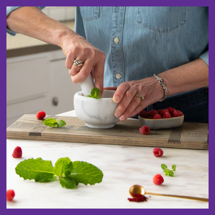 In a denim shirt, someone uses a mortar and pestle to crush mint leaves for healthy aging. Fresh raspberries and mint spread on a white counter, with more raspberries in a bowl. Nearby lies a gold spoon. Scene framed with purple, highlighting New Chapter’s Every Woman™s One Daily 55+ Multivitamin.