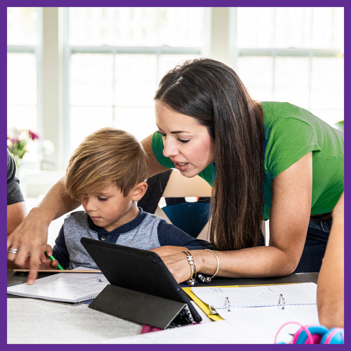 A woman with long dark hair helps a blond boy, both focused on a tablet. Open binders and papers surround them in a bright room. They feel energized and supported as if they just took New Chapters Every Woman™s One Daily Multivitamin, which supports immune health, energy, stress relief, and beauty.