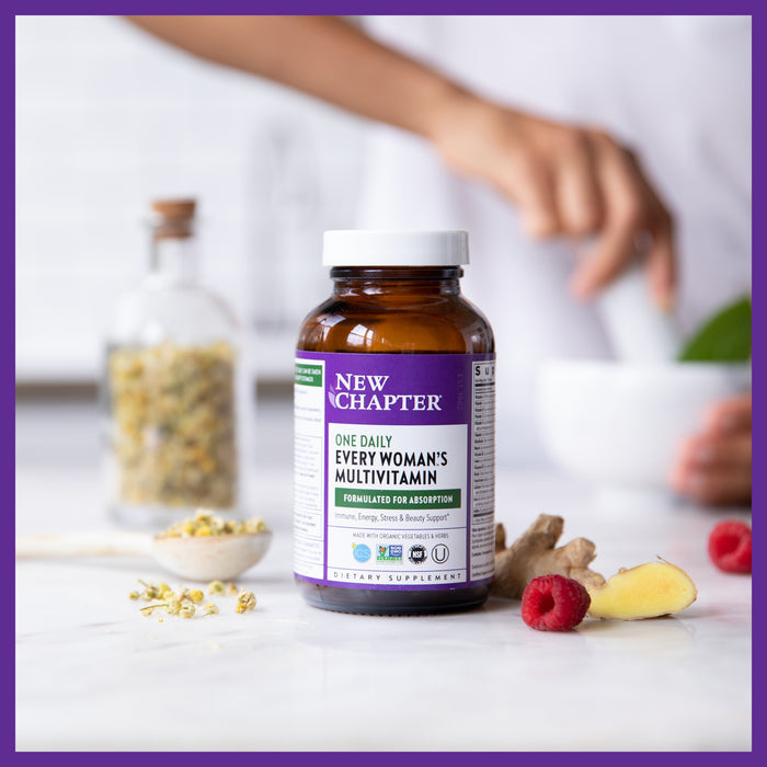 On a kitchen counter, a bottle of New Chapter Every Woman™s One Daily Multivitamin offers essential nutrients with probiotics for immune support. Nearby are fresh raspberries, ginger, and herbs as a blurred figure prepares ingredients in the background.
