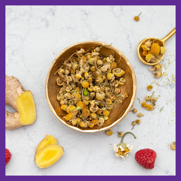 A bowl of dried chamomile flowers sits on a marble surface with sliced ginger, a raspberry, and a golden spoon of chamomile nearby—offering natural support. The image is elegantly framed with a purple border, echoing New Chapters Every Woman™s One Daily Multivitamin for immune, energy & beauty.