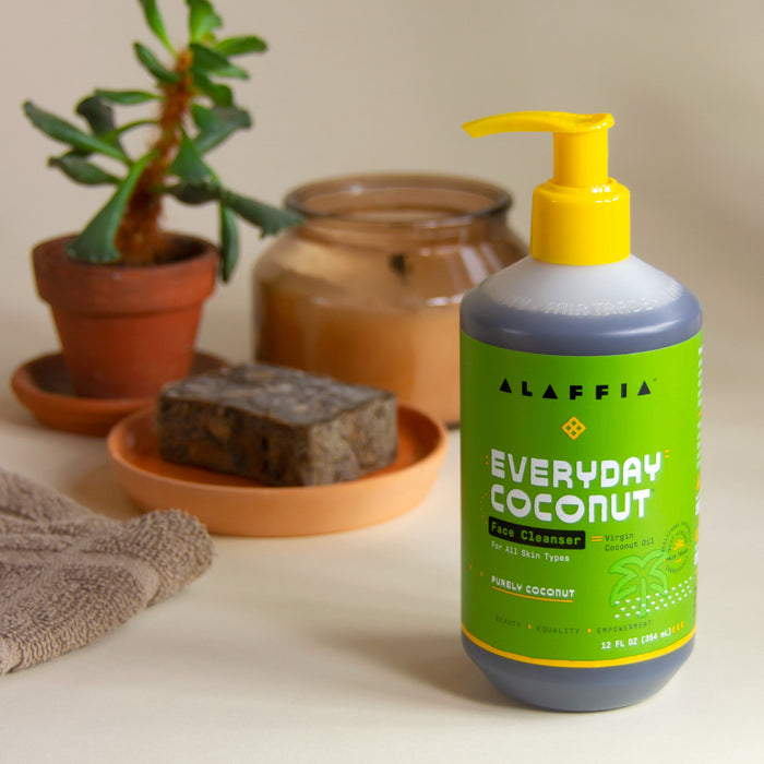 An Alaffia Everyday Coconut Face Cleanser with a yellow pump sits next to a plant in a terracotta pot, soap on a dish, and a folded towel, crafting a soothing bathroom ambiance. Its sulfate-free formula and fair trade origins enhance its appeal.