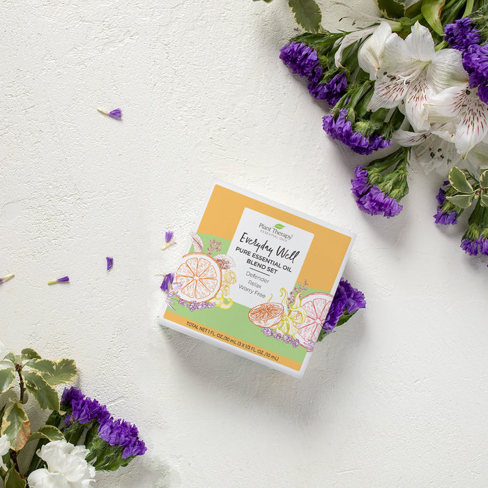 A square box of Everyday Well Pure Essential Oil Blend Trio by Plant Therapy sits on a textured white surface, surrounded by scattered purple and white flowers. The immune-boosting blends packaging features citrus illustrations in a refreshing yellow and white design.