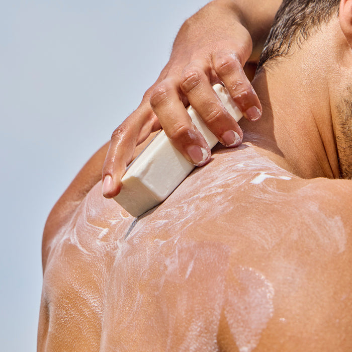 A person washes their back with Phil’s Everything Bar, a white shampoo bar enriched with chia seed oil. The close-up shows their hand, the nourishing bar, and a rich lather on their upper back.