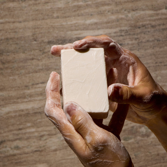 Two hands lather the rectangular white Everything Bar by Phil's, enriched with upcycled plant squalane, against a beige textured background.