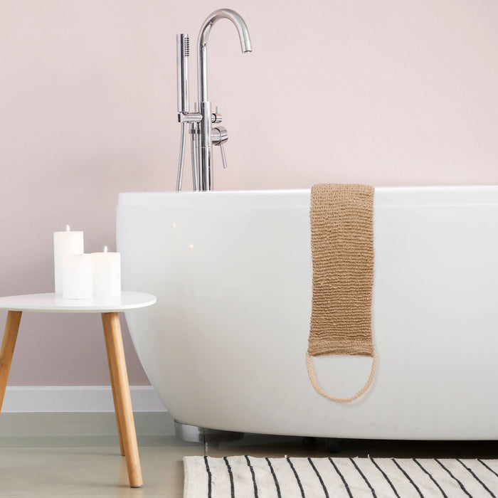 A modern bathroom showcases a white freestanding bathtub with an EcoTools Exfoliating Back Scrubber draped over the side, complemented by a woven bath mat. Nearby, a small wooden table holds three lit candles against a beige wall and striped rug.
