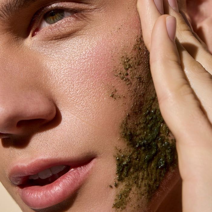 A close-up of someone applying Phil's Exfoliating Facial Cleanser with French green clay to their cheek, showcasing its textured formula for smooth skin in natural light.
