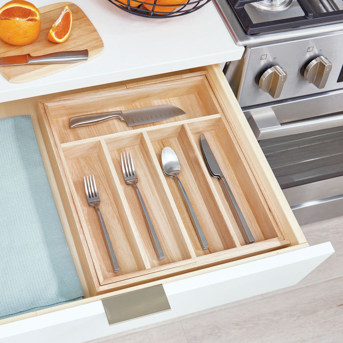 Open the kitchen drawer to reveal the iDesign Expandable Wood Cutlery Tray, holding a chefs knife, three forks, a spoon, and a butter knife. A light blue cloth is visible on the left, with a stove on the countertop to the right.