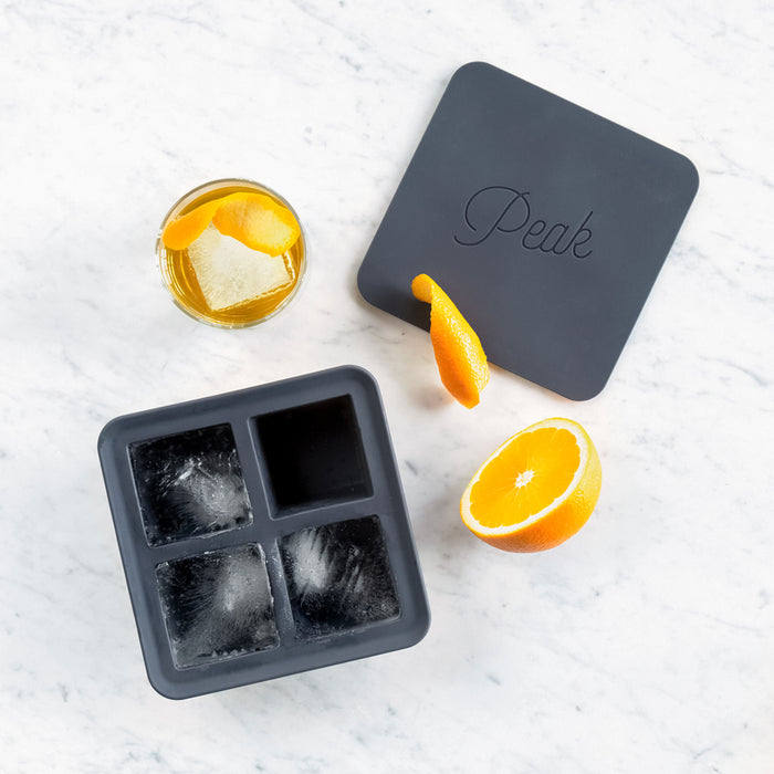 The Extra Large Ice Cube Tray by W&P sits on a white marble surface. Nearby are half an orange, a drink with slow-melting ice, an orange peel twist, and a black lid labeled Peak.