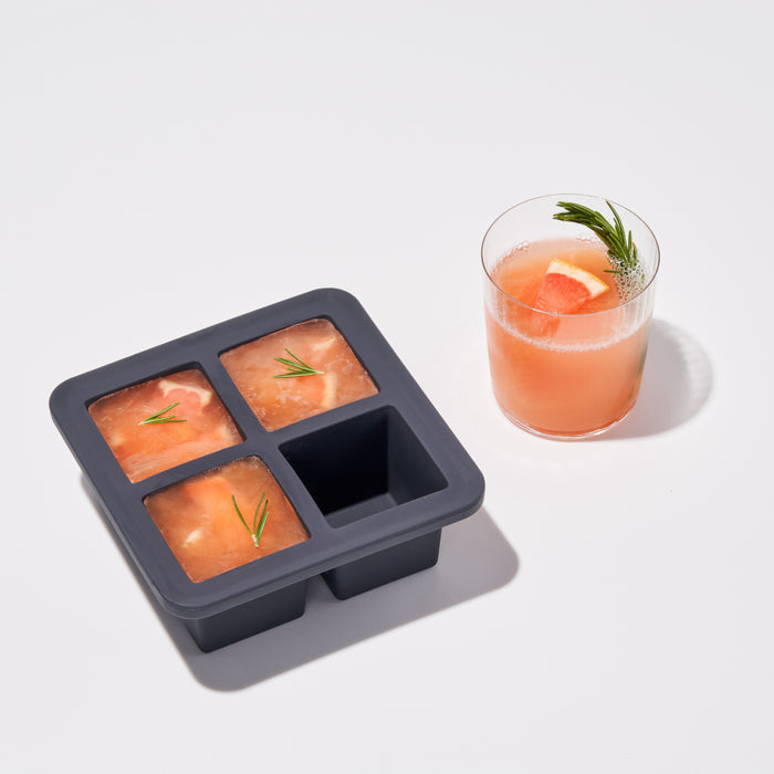 A pink drink with citrus and dill sits next to W&Ps Extra Large Ice Cube Tray, which holds slow-melting pink cubes topped with dill sprigs, all on a light surface.