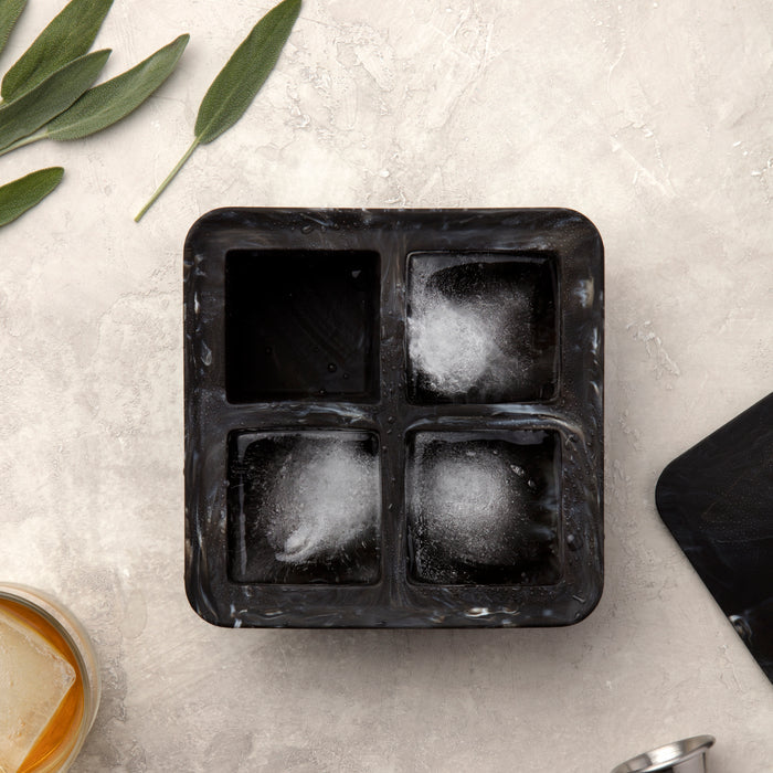 The W&P Extra Large Ice Cube Tray, black with four compartments, partially filled and forming ice crystals, is designed for perfect portion sizes. It rests on a light textured surface with green leaves and a glass of iced drink in the corner.