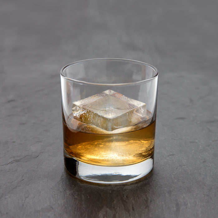 A glass of whiskey with a slow-melting W&P Extra Large Ice Cube rests on a dark, textured surface. The whiskeys golden-brown hue shimmers, reflecting off the ice and glass for a polished, inviting look.