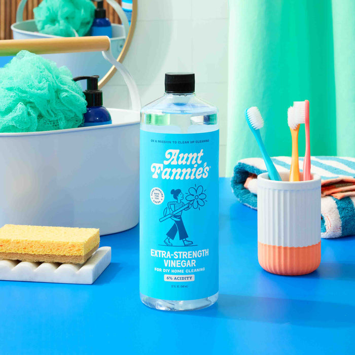 A bottle of Aunt Fannies Extra Strength Vinegar: For Cleaning rests on a blue bathroom counter with two toothbrushes, a sponge, washcloth, soap dispenser, and green bath pouf. A turquoise curtain is visible in the background.