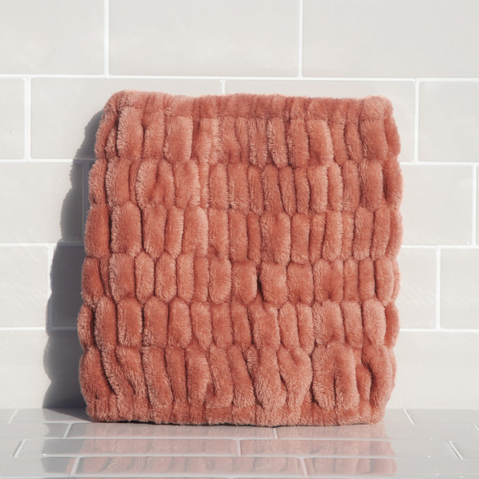 A textured, rust-colored, plush square cushion sits against a white tiled wall. It has a grid pattern with raised sections for a cozy look. Sunlight casts subtle shadows similar to those on Kitschs Extra Wide Spa Headband, enhancing the cushions texture.