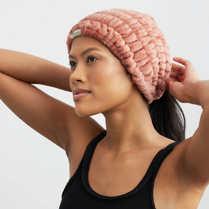 A person wearing Kitschs eco-friendly Extra Wide Spa Headband in pink and a black sleeveless top poses with arms lifted behind their head against a plain, light-colored background, adding simple elegance to their hair care routine.