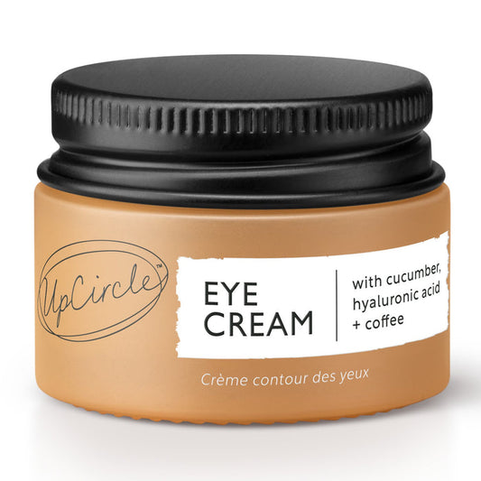 The UpCircle Eye Cream, designed to reduce puffiness and dark circles, features a circular handwritten logo on its beige label that says Eye Cream with Cucumber, Hyaluronic Acid + Coffee and Crème contour des yeux. It comes in a small jar with a black lid.