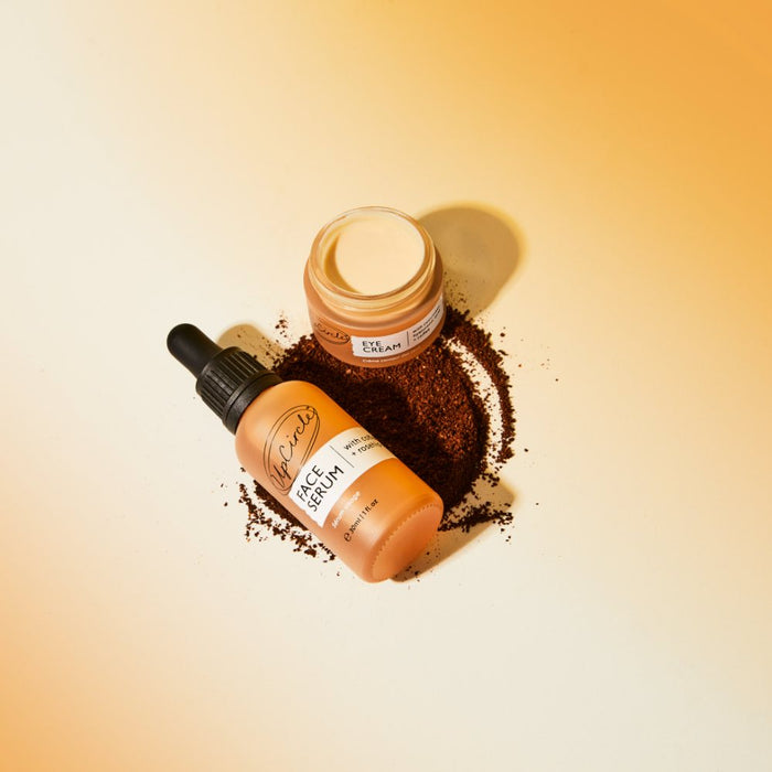 An UpCircle Eye Cream jar with cucumber, hyaluronic acid, and coffee sits alongside a face serum bottle featuring droppers on a light orange surface. Coffee grounds, known for reducing puffiness and dark circles, form a textured circular pattern around bottles with orange and brown labels.