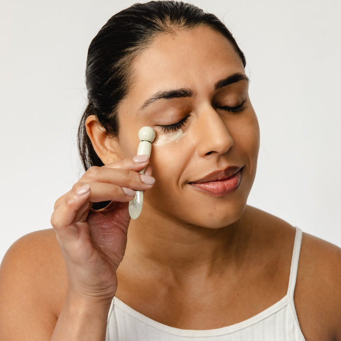 A dark-haired woman in a white sleeveless top smiles gently with closed eyes as she applies UpCircles Eye Cream with Cucumber, Hyaluronic Acid + Coffee to her right eye area using an applicator, targeting puffiness and dark circles against a plain white background.