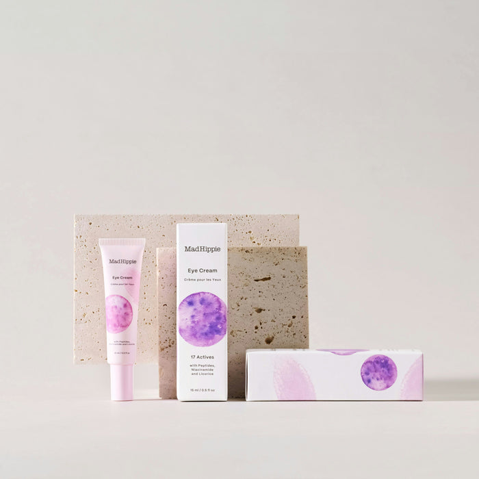 A tube and box of Mad Hippie Eye Cream, featuring antioxidant-rich benefits, are set against a minimalist backdrop. The packaging displays a purple watercolor design with product details. Textured beige rectangular blocks are nearby.